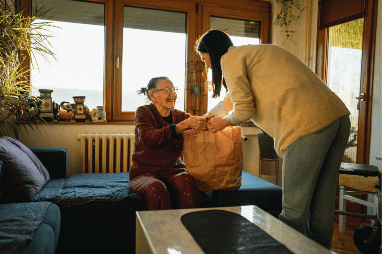 A woman in a sweater provides home care by handing a paper bag to a smiling older woman sitting on a couch. Our home care cost model shows the true cost of care.