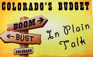 Wooden signposts depicting a metaphor for Colorado's economic status with arrows pointing in opposite directions labeled "boom" and "bust," under the title "Colorado Budget Explanation in Plain Talk.