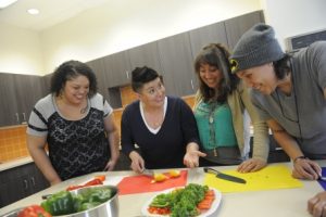 Four individuals smiling and engaged in a unique cooking activity around a kitchen counter with fresh vegetables.