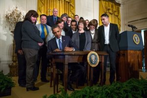 President signing an overtime pay bill into law at a desk with onlookers observing the ceremony.