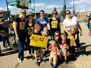 Group of people, including children, holding a sign advocating for $12 minimum wage, gathered outdoors on a sunny day in 2016 highlights.