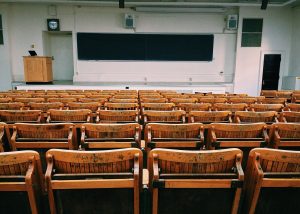 An empty lecture hall with rows of wooden seats, higher tuition costs noted on a blackboard at the front.