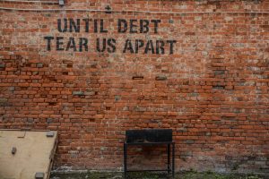 A weathered brick wall with the phrase "until debt tear us apart" stenciled on it above a lone, empty bench, hinting at the importance of secure savings.