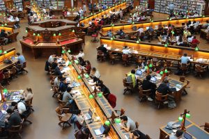 Students studying in a large, well-lit library with wooden tables and shelves filled with books, many of whom are Post-Traditional Adult Students.