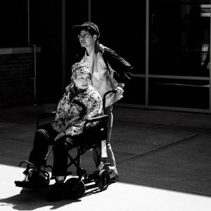 A person providing Golden Year Caregiving by pushing an elderly individual in a motorized wheelchair outdoors in a sunny setting.
