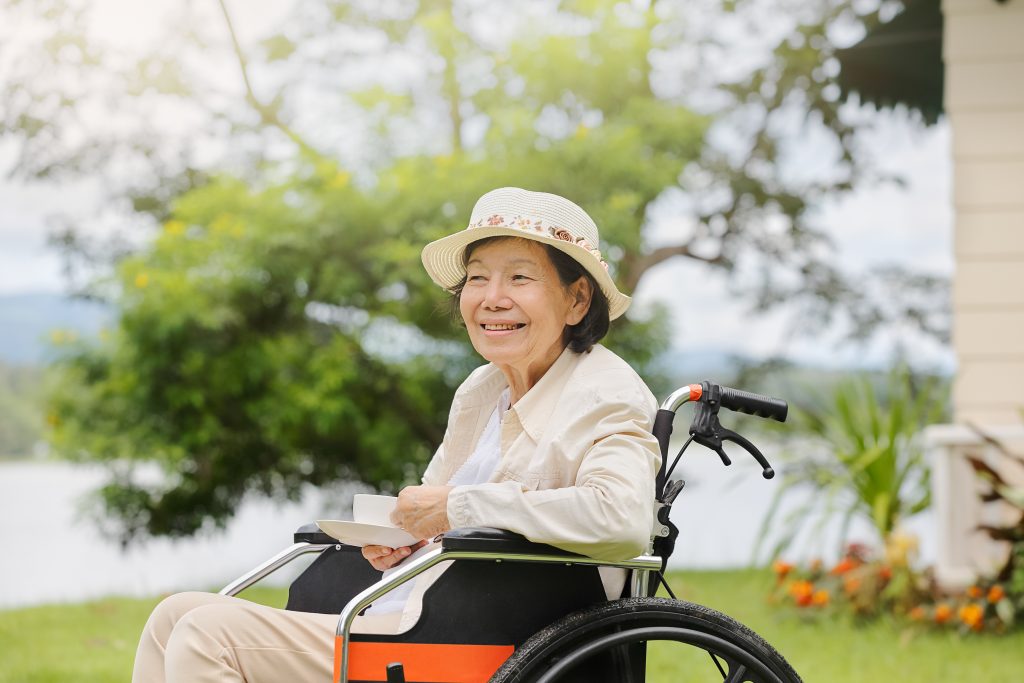 Senior woman with a hat sitting in a wheelchair outdoors, smiling with a notebook on her lap, embodying Aging in Colorado.