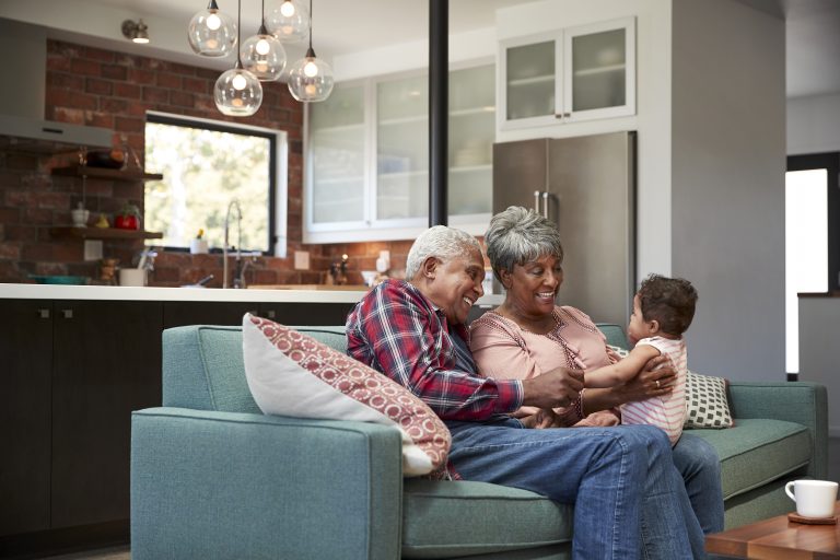 Senior couple sitting on a sofa laughing with a toddler in a modern kitchen setting, embodying the joy highlighted in the "Roadmap for Aging" in Colorado.