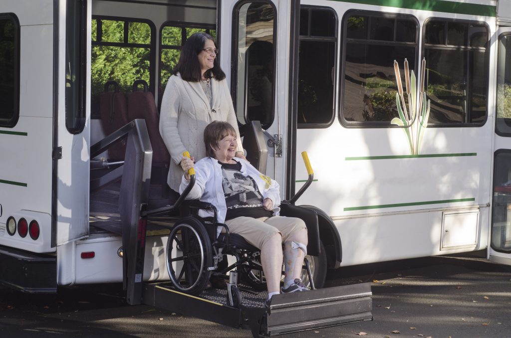 A woman in a wheelchair smiles as she is assisted by another person while using a wheelchair lift to board a bus, benefiting from Colorado's Elderly Care Services.