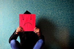 Person holding a red paper with a sad face in front of their head against a textured wall, symbolizing the student debt scale.