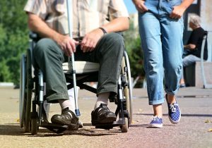 A person in a wheelchair accompanied by someone walking beside them on a sunny day.