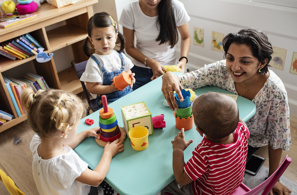 Adult supervising three young children playing with toys at a round table as part of Colorado's Early Childhood Education workforce.