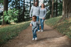 A young child walks ahead on a dirt path while two adults discuss policy proposals follow behind, amidst a backdrop of trees.