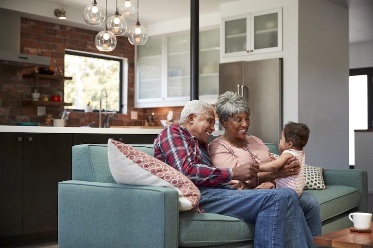 A senior couple sitting on a sofa in a modern kitchen living area, smiling and playing with a young family member.