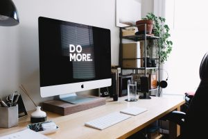 A modern, tidy workspace with an iMac displaying the motivational message "do more," surrounded by office supplies, a potted plant, and documents concerning Colorado policy.