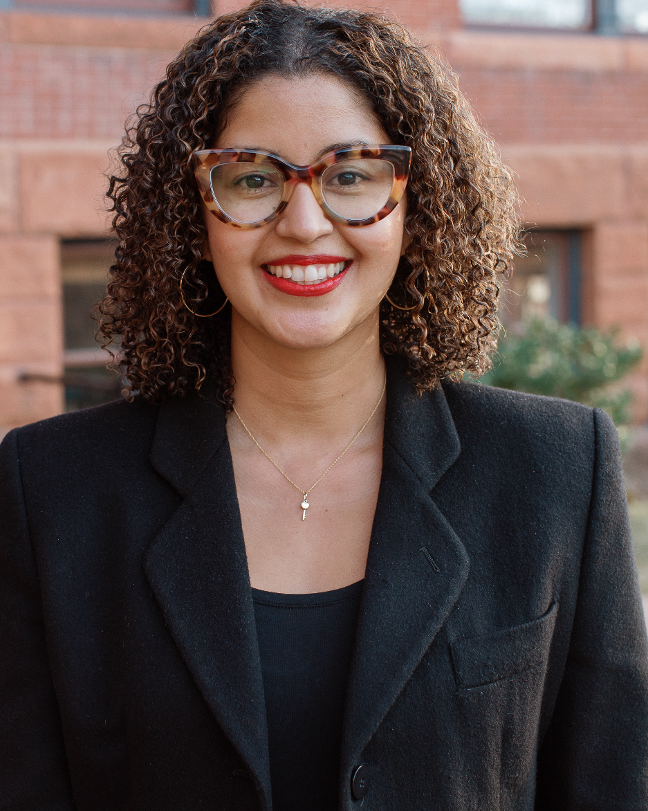 A smiling woman with curly hair wearing glasses and attending a board meeting in a black coat.