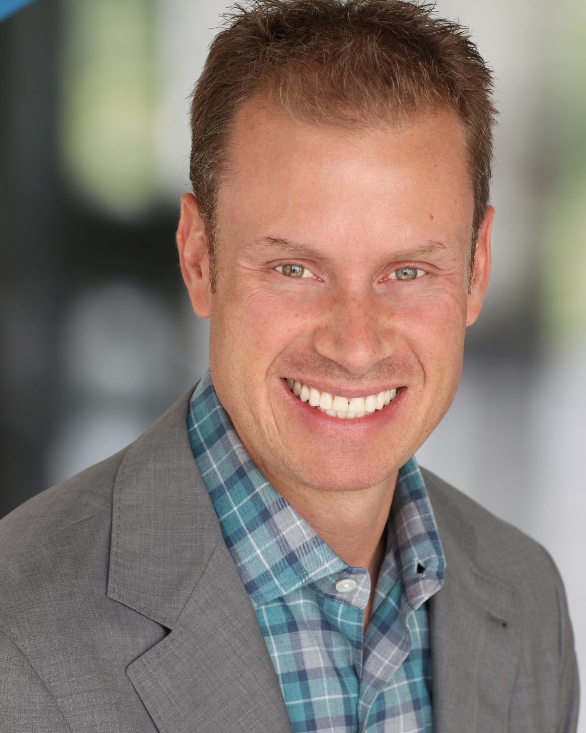 A smiling man with short blond hair, wearing a grey blazer over a checkered shirt, looks like he could be among the board members.