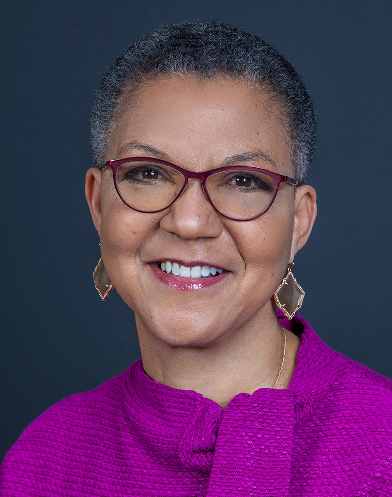 A smiling woman with short gray hair and red glasses, celebrating her 20th anniversary, wearing a purple sweater and gold earrings, against a dark background.