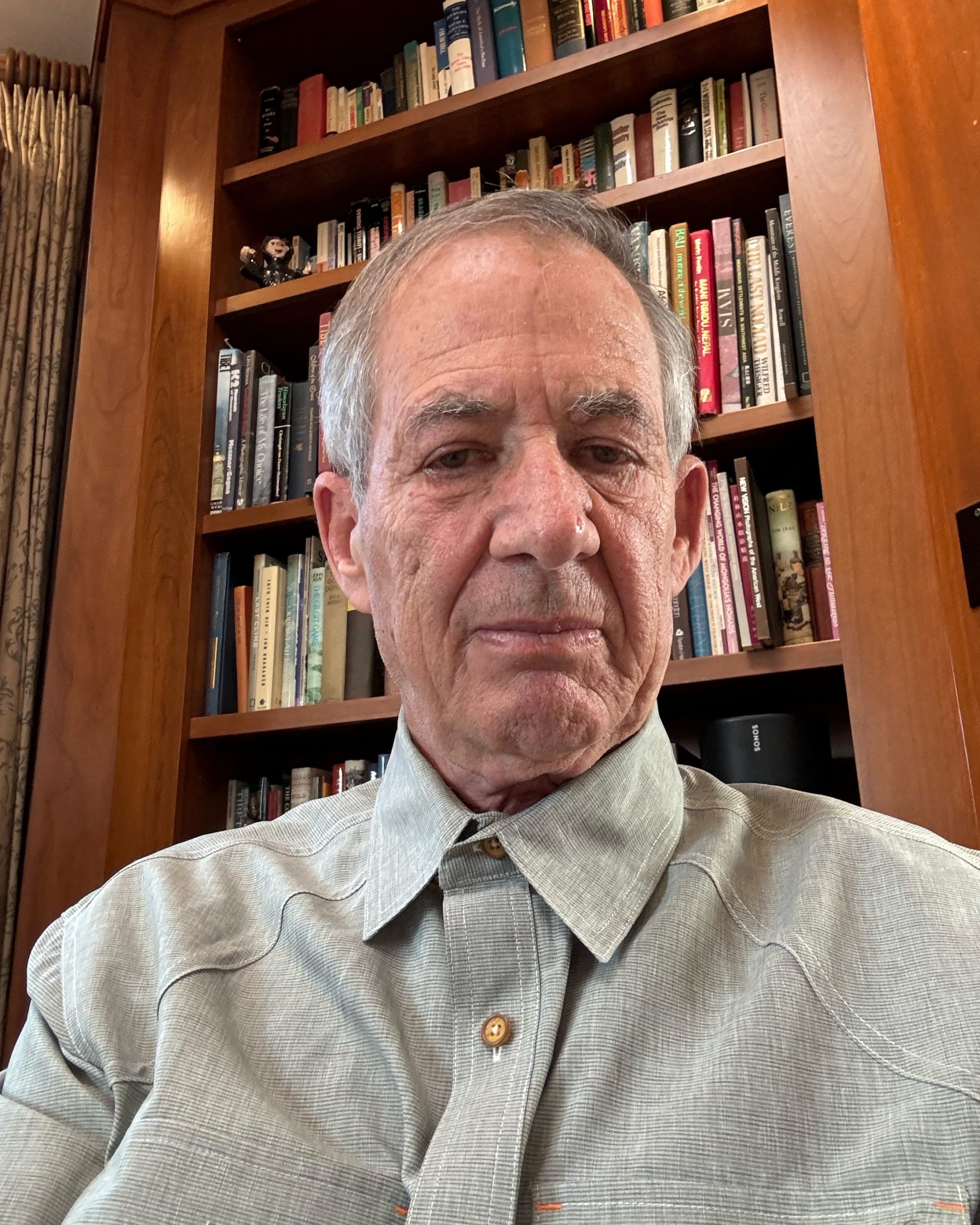 Elderly man in a gray shirt sitting in front of a board and bookshelf.