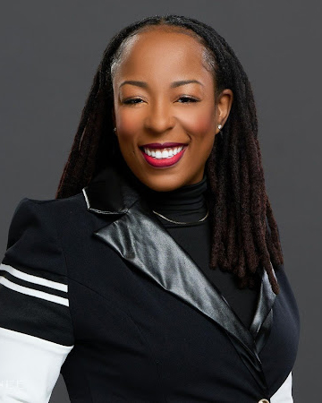 A woman with dreadlocks smiling at the camera, wearing a black outfit with leather accents, white stripes, and a board-inspired design.