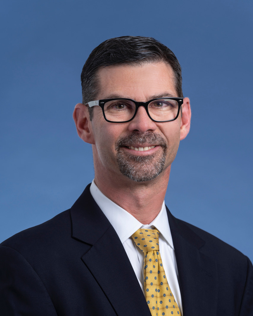 Portrait of a smiling man with glasses, wearing a dark suit and a yellow patterned tie against a blue background, suggesting he could be part of management board.