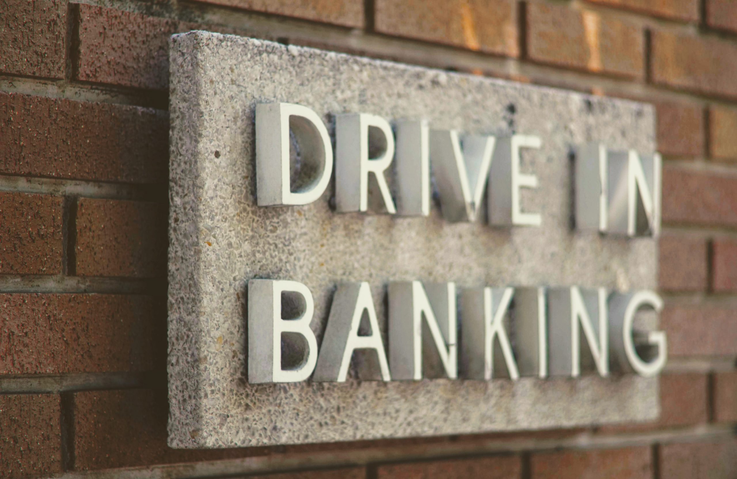 Sign indicating 'drive in banking options' mounted on a brick wall.